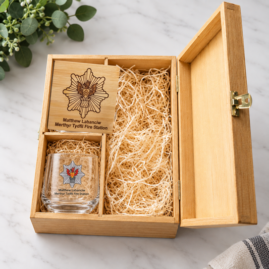 A personalised wooden gift box with an engraved fire station emblem and name, containing a clear glass tumbler with a matching fire service crest and shredded paper filler, photographed on a light marble surface.