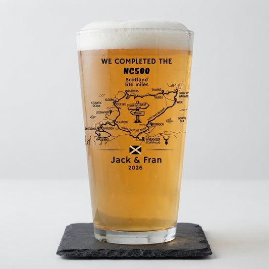 A pint glass filled with golden beer, standing on a dark slate coaster against a light gray background. The glass is printed with a black map of Scotland, the text 'WE COMPLETED THE NC500 Scotland 516 miles', the names 'Jack & Fran', and the year '2026', along with a small black saltire flag.