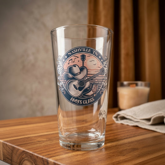 A clear glass pint glass with a printed Nashville Tennessee 'Amy's Glass' graphic featuring a cowboy hat and guitar, set on a wooden table with a blurred background.
