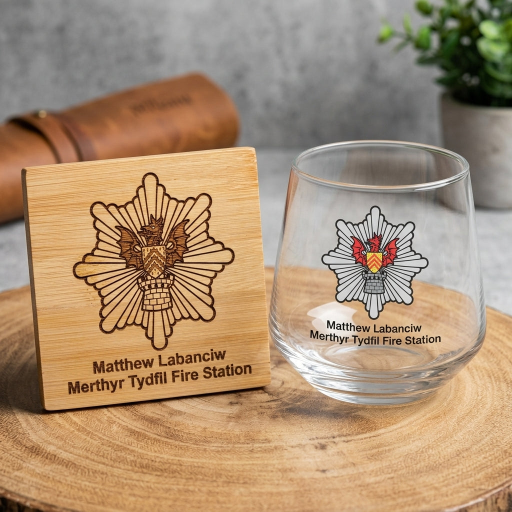 A clear stemless wine glass and a square wooden coaster engraved with the Merthyr Tydful Fire Station emblem and the name Matthew Labanciv, displayed on a round wooden slice with a blurred background.
