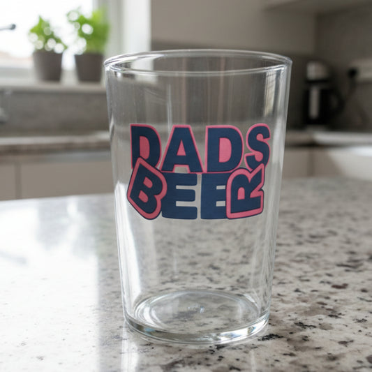 'Dads Beer' pint glass featuring bold red and orange lettering design, filled with golden ale, set on kitchen counter with blurred home background