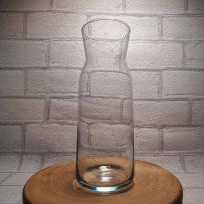 A clear glass carafe with no contents, displayed on a wooden surface against a white brick wall background.