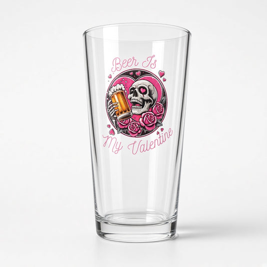 A clear pint glass with a pink and black graphic design featuring a skull holding a beer mug, surrounded by roses, with the text 'Beer Is My Valentine' in pink script.