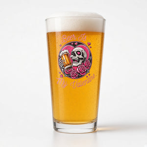 A pint glass filled with golden beer and topped with white foam, featuring a colorful graphic of a skull holding a beer mug surrounded by pink roses and the text 'Beer Is My Valentine'.