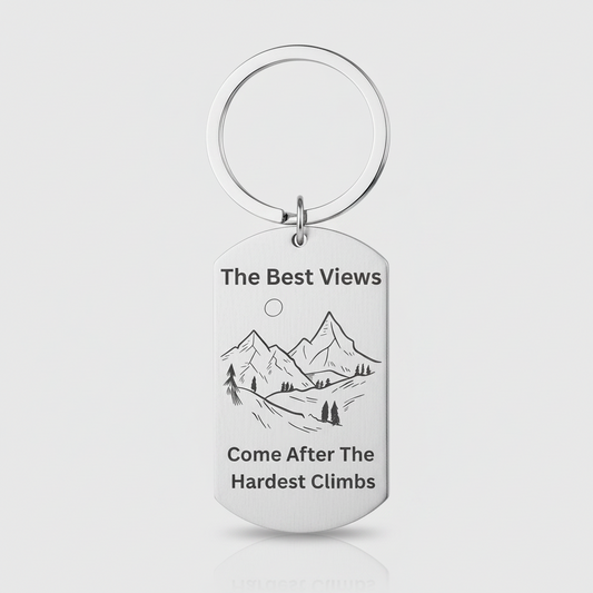 A silver metal dog tag keychain with a circular split ring, featuring a black engraved mountain illustration and the text 'The Best Views Come After The Hardest Climb' on a light gray background.