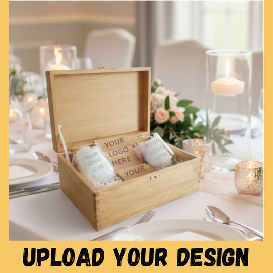 Open wooden keepsake box containing two personalised engraved stemless glasses with custom logo, displayed on a wedding reception table with candles and blush roses in the background