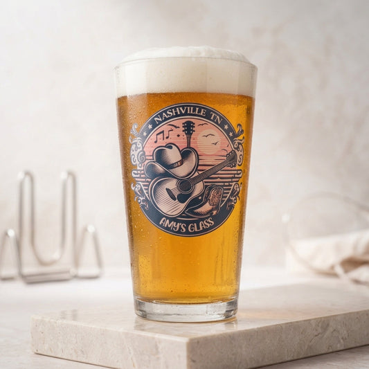 A pint glass filled with golden beer and topped with white foam, standing on a light stone surface. The glass has a circular printed design featuring a cowboy playing guitar with the text 'Nashville in Emmy's Class'.