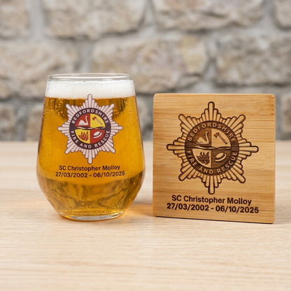 A clear stemless beer glass filled with golden beer and a foam head, engraved with the Fire and Rescue Service crest and personal details, stands beside a matching wooden coaster on a light wood surface with a blurred stone wall background.