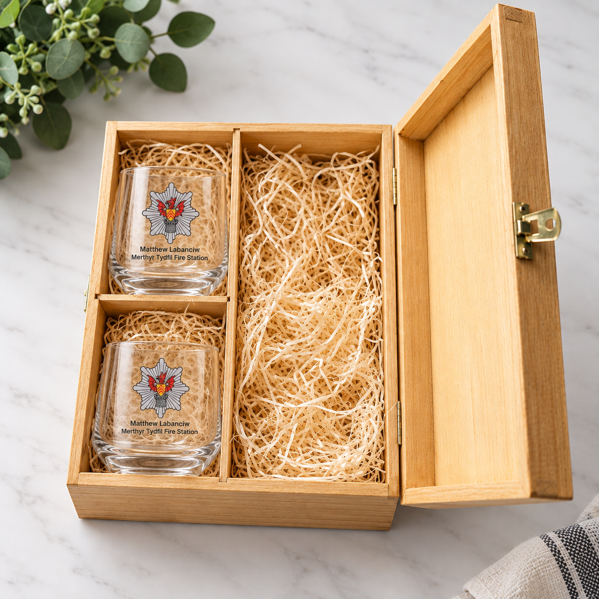 A wooden gift box with an open lid, filled with shredded paper and holding two clear glass tumblers printed with a fire department emblem and the text 'Matthew Roberts Middleburg Fire Department.' The box is photographed on a light marble surface with a small plant and fabric visible at the edges.