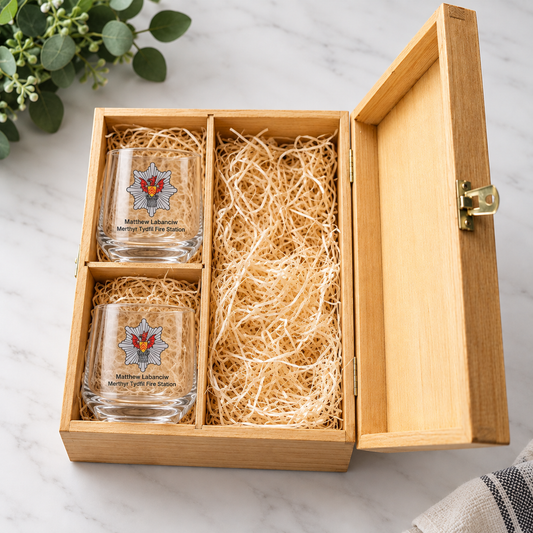 A wooden gift box with an open lid, filled with shredded paper and holding two clear glass tumblers printed with a fire department emblem and the text 'Matthew Roberts Middleburg Fire Department.' The box is photographed on a light marble surface with a small plant and fabric visible at the edges.
