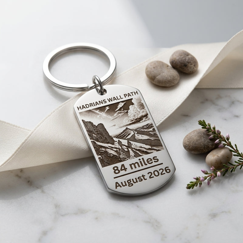 A personalised silver metal keychain with an engraved dog tag featuring a landscape illustration and the text 'HADRIANS WALL PATH', '84 miles', and 'August 2026'. The keychain is styled on a cream ribbon over a white marble surface with decorative stones and a sprig of purple flowers.