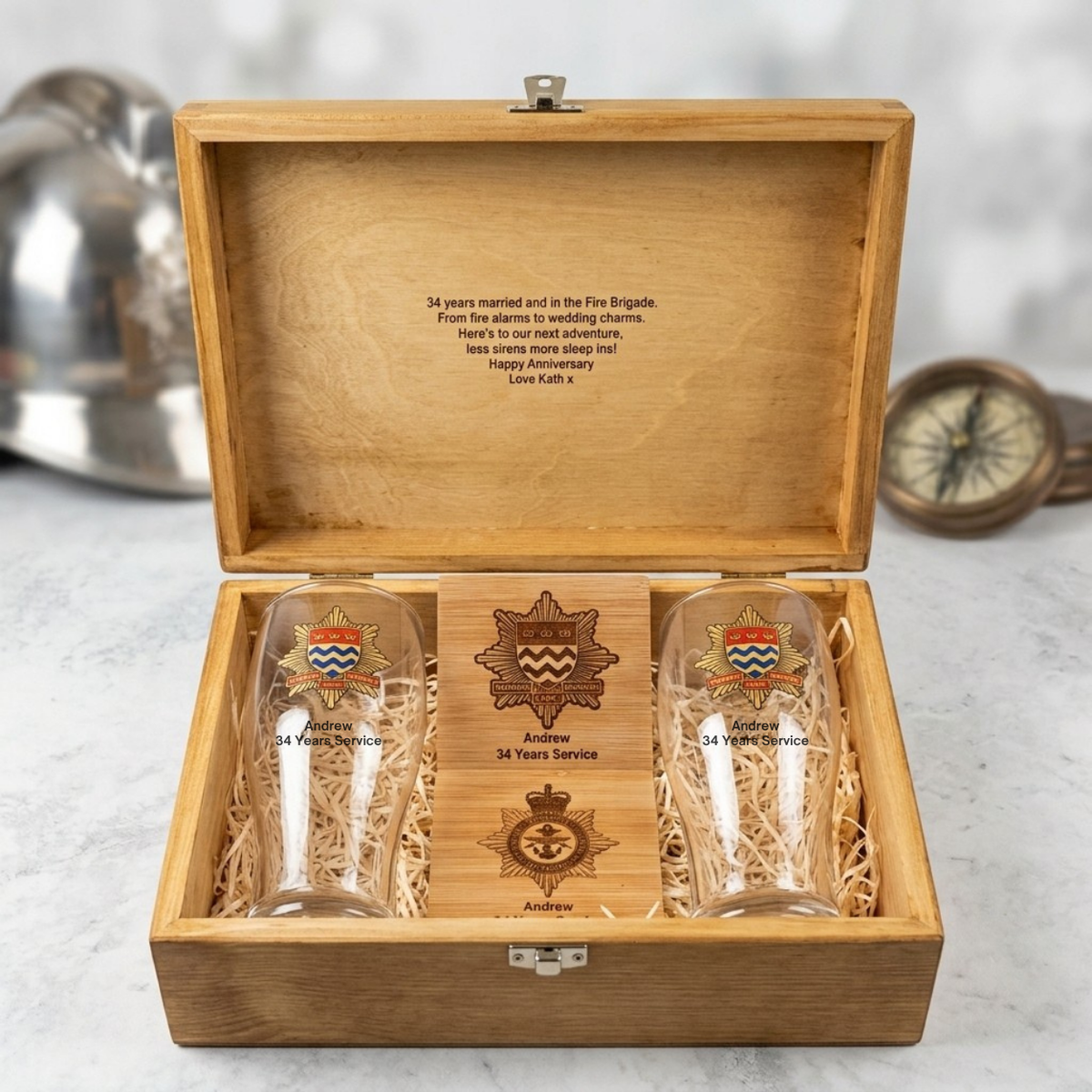 A wooden presentation box with the lid open, containing two clear engraved beer glasses and a central wooden plaque with military-style insignia. The box is placed on a light marble-like surface with blurred metal and wood objects in the background.