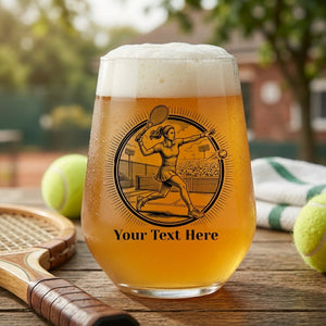a stemless glass with a female tennis player design and custom text resting on a table with a tennis racket and tennis balls 