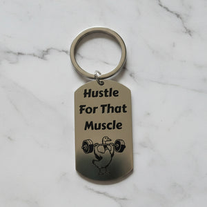 Keychain with 'Hustle For That Muscle' text and graphic on a light grey background