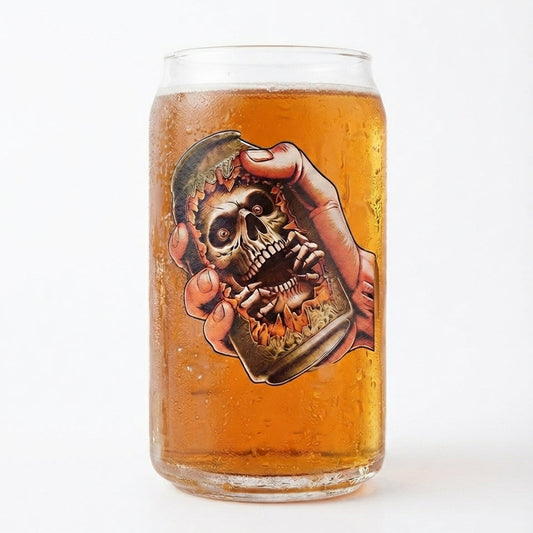 A clear glass can-shaped mug filled with amber beer, featuring a large graphic of a skull held by a hand on the front, set against a plain white background.