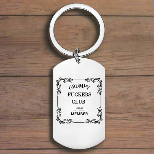 A silver keyring with the phrase 'GRUMPY FUCKERS CLUB LIFETIME MEMBER' printed in bold letters, set against a wooden background.