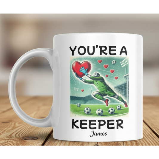 A white ceramic mug with a custom graphic print featuring a soccer goalkeeper diving to save a red heart, with the text 'YOU'RE A KEEPER James' on a wooden table with a blurred background.
