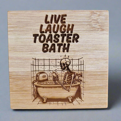 Laser engraved bamboo coaster with “Live Laugh Toaster Bath” text and skeleton in bathtub with toaster – dark humor eco-friendly gift, goth or emo room décor
