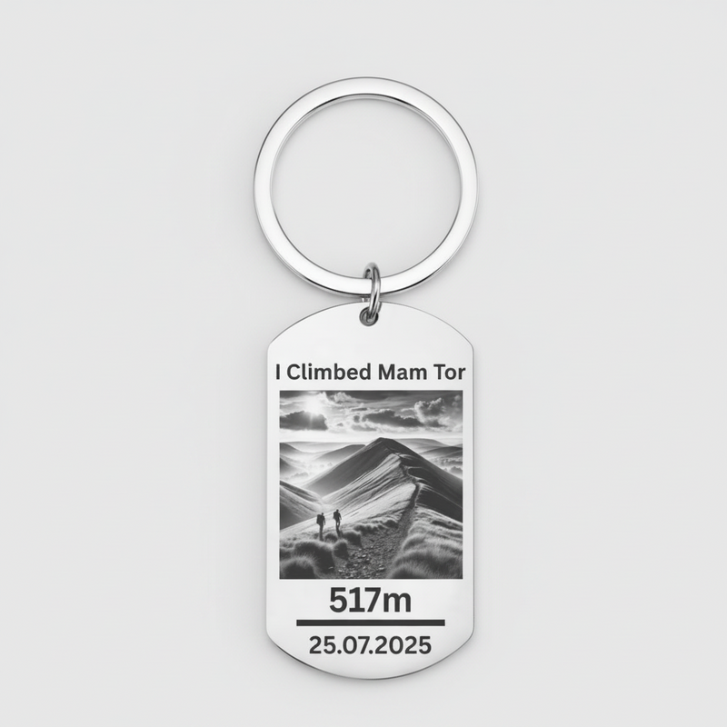 A silver metal dog tag keychain with a circular key ring, featuring a black and white photo of a mountain landscape with the text 'I Climbed Mam Tor', '517m', and '25.07.