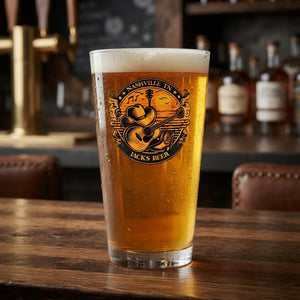 A frosted pint glass filled with golden beer, featuring a circular Nashville Tennessee 'Jacks Beer' logo, set on a rustic wooden bar table with blurred bottles and taps in the background.
