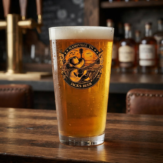 A frosted pint glass filled with golden beer, featuring a circular Nashville Tennessee 'Jacks Beer' logo, set on a rustic wooden bar table with blurred bottles and taps in the background.