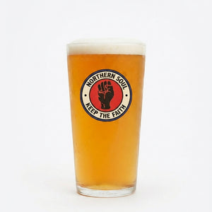 A pint glass filled with amber beer and topped with white foam, featuring a circular Northern Soul 'Keep The Faith' logo on the front, set against a plain white background.