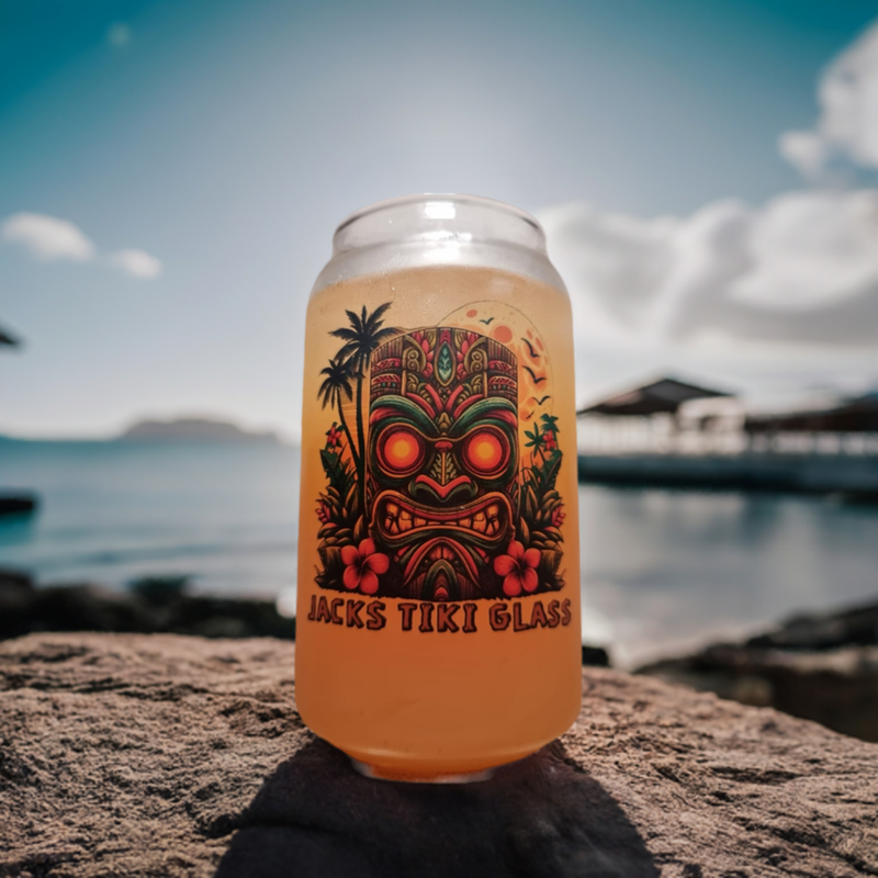 A personalised tiki can-shaped cocktail glass with a colorful tiki mask and floral design, placed on a rock with a tropical background.