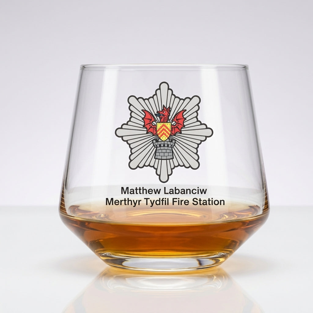 A clear stemless whiskey glass with an amber liquid inside, featuring a printed Merthyr Tydfil Fire Station emblem and the names Matthew Labanciw and Merthyr Tydfil Fire Station on a white background.