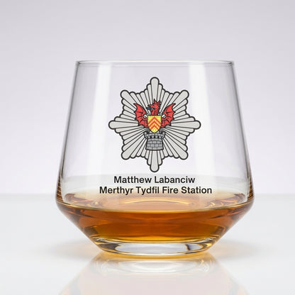 A clear stemless whiskey glass with an amber liquid inside, featuring a printed Merthyr Tydfil Fire Station emblem and the names Matthew Labanciw and Merthyr Tydfil Fire Station on a white background.