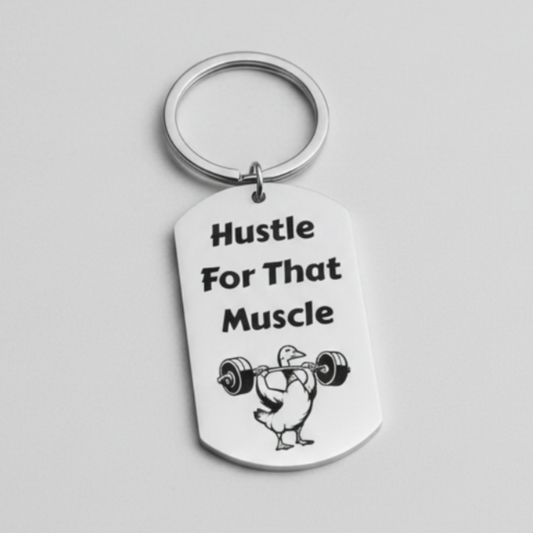 A silver metal dog tag keychain with a black engraved design that reads 'Hustle For That Muscle' alongside a cartoon illustration of a person lifting a barbell, set against a plain light gray background.