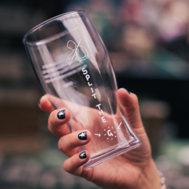 A hand with black nail art holds a clear, tapered glass tumbler with white printed text and graphics. The background is a heavily blurred, dark, out-of-focus environment.