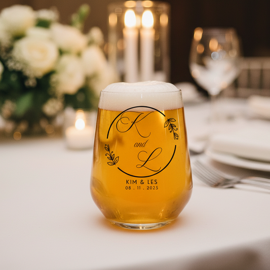Personalised stemless glass printed with a botanical monogram design reading K and L, Kim and Les, 08.11.2025, filled with beer on a white wedding reception table with candles and white roses in the background