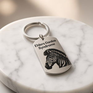 A silver metal dog tag keychain engraved with the text 'Ehlers Danlos Syndrome' and a black zebra illustration, resting on a white marble coaster.