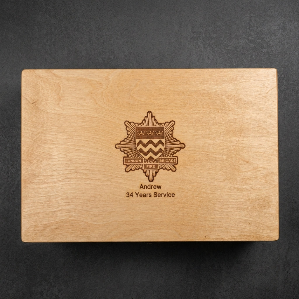 A rectangular wooden keepsake box lid with a natural light wood grain finish, featuring an engraved military-style crest and the text 'Andrew 34 Years Service' in a darker brown tone, set against a dark textured background.