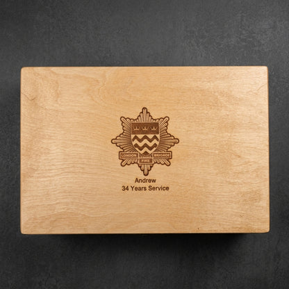 A rectangular wooden keepsake box lid with a natural light wood grain finish, featuring an engraved military-style crest and the text 'Andrew 34 Years Service' in a darker brown tone, set against a dark textured background.