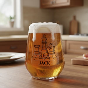 A clear stemless wine glass filled with golden beer and topped with white foam, printed with a black chess-piece graphic and the name "JACK," set on a wooden kitchen table.