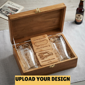 An open wooden gift box on a light gray surface, containing two clear engraved beer steins and a personalized wooden plaque. A beer bottle and a magazine are visible in the background as props.