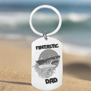 Fintastic Dad laser engraved stainless steel dog tag keyring featuring bold shark illustration, photographed on sandy beach background, funny personalised Father's Day gift for fishing dads