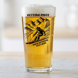A clear pint glass filled with golden beer and topped with white foam, standing on a light surface. The glass has a black printed graphic of a skier in front of mountains, with the text 'GETTING PISTE' at the top and 'YOUR OPTIONAL TEXT HERE' below.