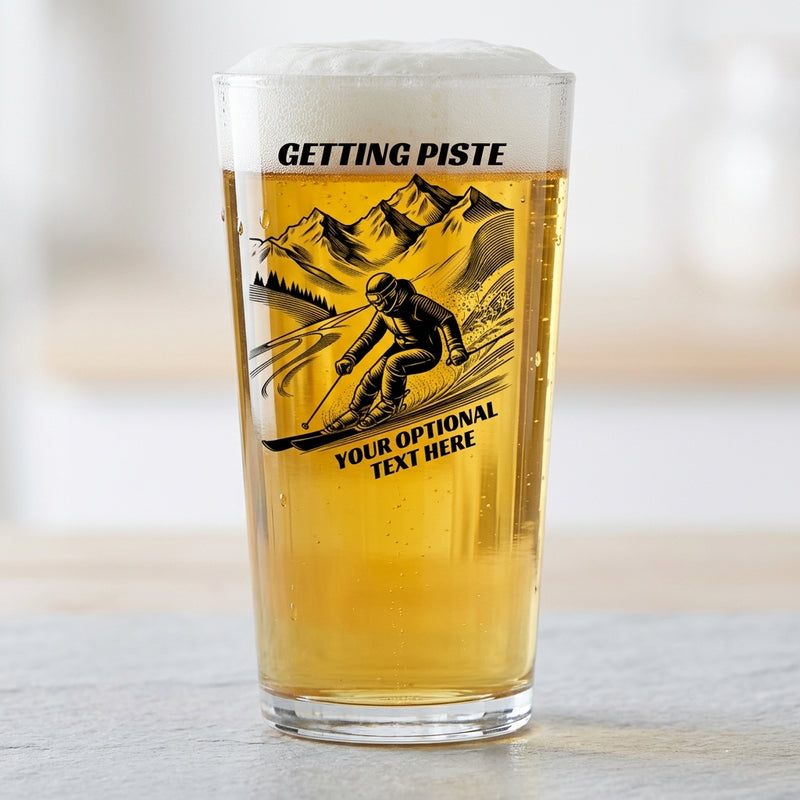 A clear pint glass filled with golden beer and topped with white foam, standing on a light surface. The glass has a black printed graphic of a skier in front of mountains, with the text 'GETTING PISTE' at the top and 'YOUR OPTIONAL TEXT HERE' below.