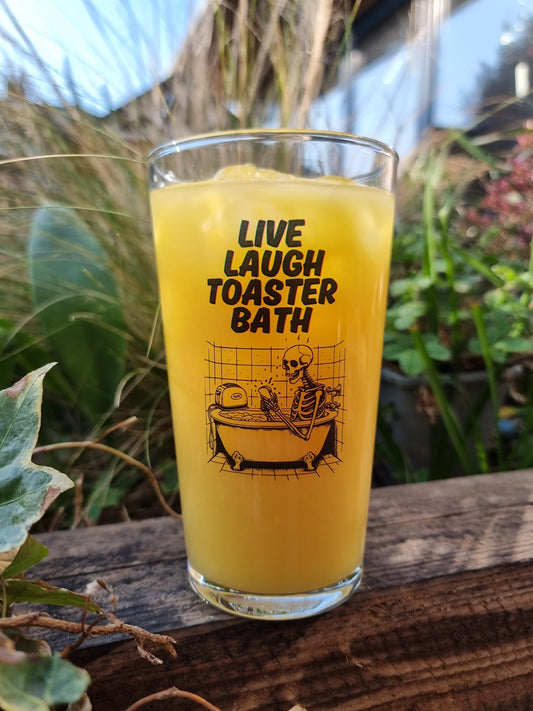 Clear drinking glass with “Live Laugh Toaster Bath” text and skeleton in bathtub with toaster – funny dark humor glass for goth, emo, or alternative gifts