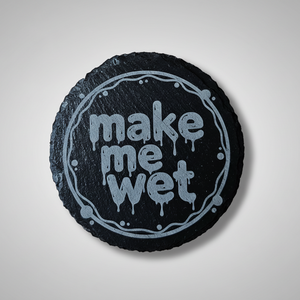 A round black slate coaster featuring the phrase 'make me wet' in a light blue dripping font, surrounded by a decorative circular border, set against a neutral gray background.