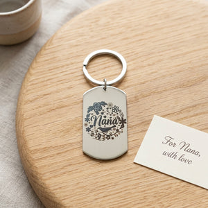 A silver metal dog tag keychain engraved with 'Nana' and floral designs, resting on a round wooden board next to a small card that reads 'For Nana, with love'.