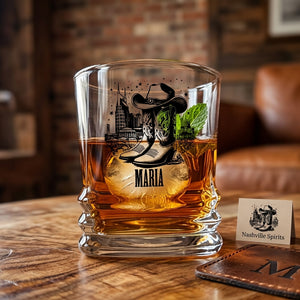 A clear whiskey tumbler with a rustic western design featuring cowboy boots, a cowboy hat, a city skyline, and the name 'MARIA' printed on it. The glass is filled with amber whiskey and a mint leaf, sitting on a wooden table next to a small 'Nashville Spirits' card.