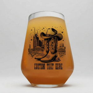 A clear stemless wine glass with a black western-themed graphic of cowboy boots and a hat, with the text 'custom text here' printed below, set on a white studio background