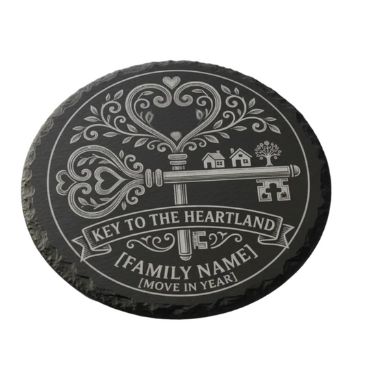 A round black slate coaster featuring a personalized engraved design with a vintage key, hearts, houses, and banners reading 'KEY TO THE HEARTLAND', '[FAMILY NAME]', and '[MOVE IN YEAR]'.