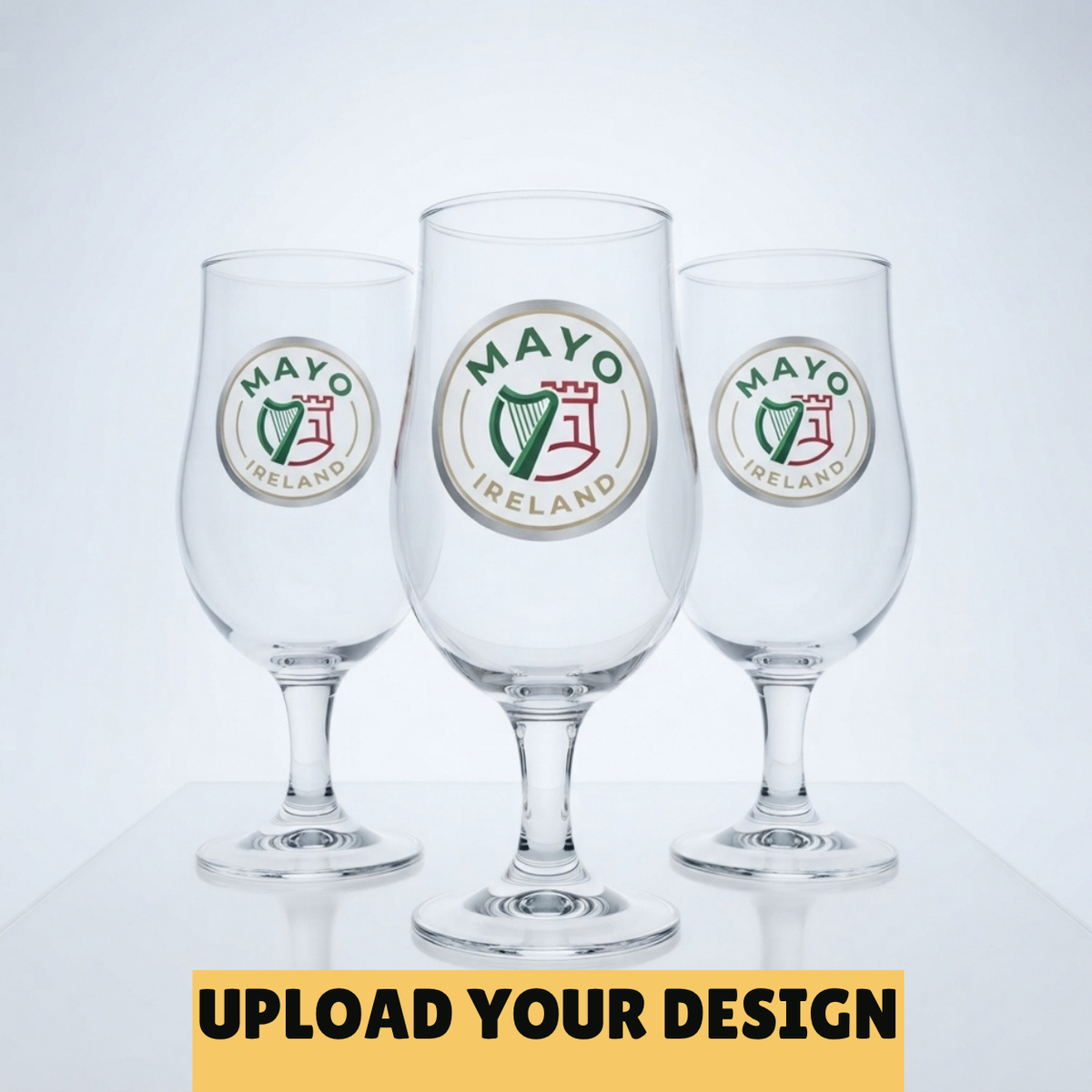 Three clear beer glasses with circular Mayo Ireland logos, shown on a light gray background with a yellow banner at the bottom reading 'UPLOAD YOUR DESIGN'.