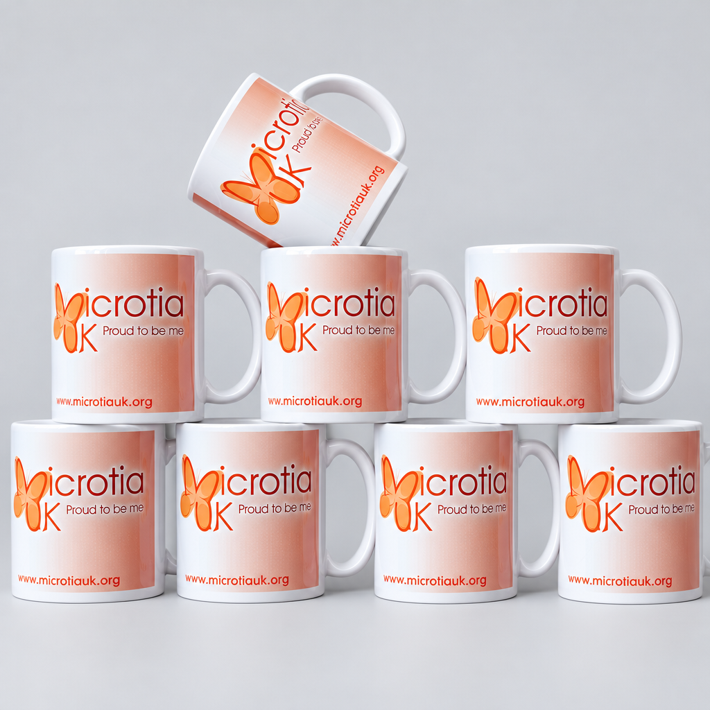 A group of white ceramic mugs printed with the Microtia K logo and website, arranged on a wooden surface against a beige stone wall. One mug is tipped on its side, and several are stacked in the foreground.