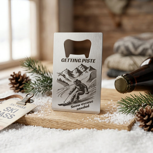 A silver metal ski-themed bottle opener with custom text, standing upright on a wooden board, surrounded by pine branches, pinecones, faux snow, a ski pass tag, and a dark bottle cap in a rustic indoor setting.