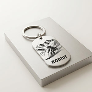 A silver metal dog tag keychain with an engraved skier and mountain design, featuring the name 'ROBBIE' in bold lettering, resting on a light gray gift box.